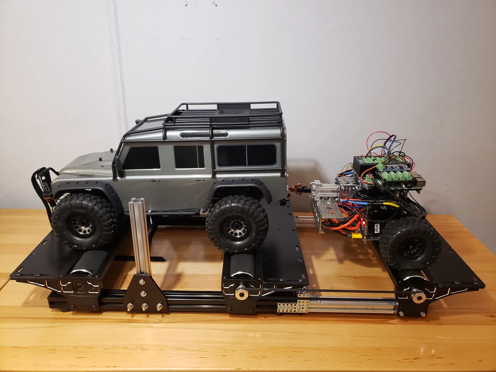 1:10 scale model SUV on a roller chassis dyno, towing an instrumented trailer robot with exposed controller boards and wiring.